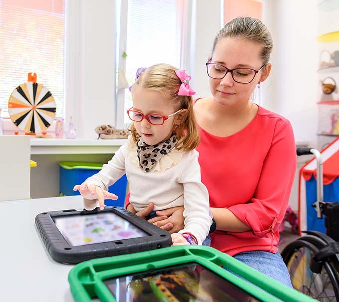 Photo of a young girl sitting on the lap of a young woman helping her with an AAC device.